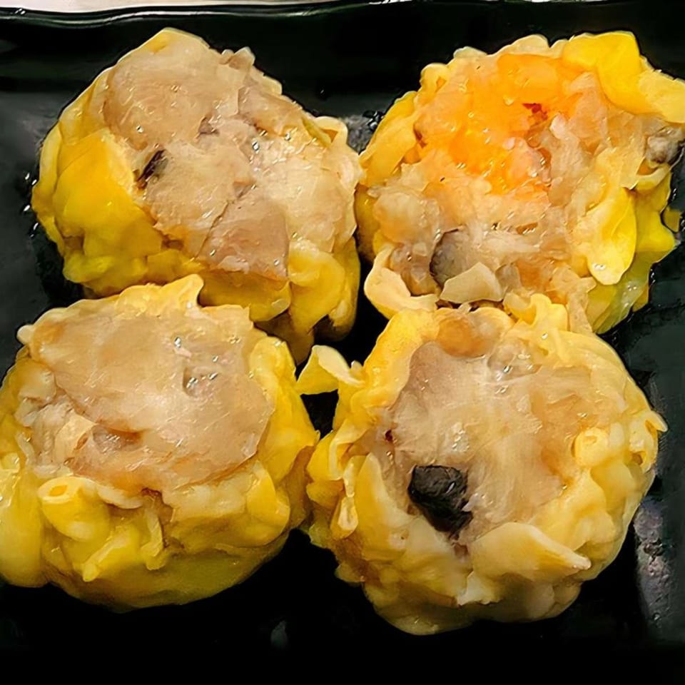 Four steamed Chinese shumai dumplings with yellow wrappers and savory filling on a black plate.