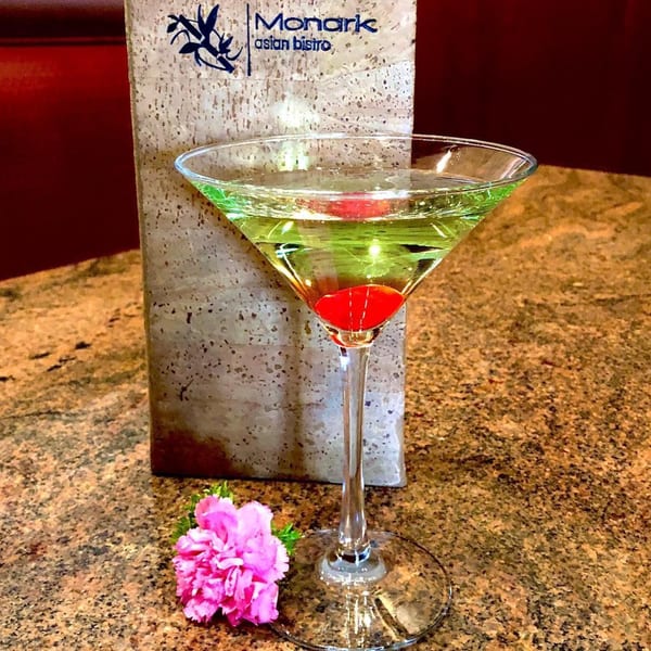 Green martini with a cherry, a pink flower, and a Monark Asian Bistro menu on a granite table.