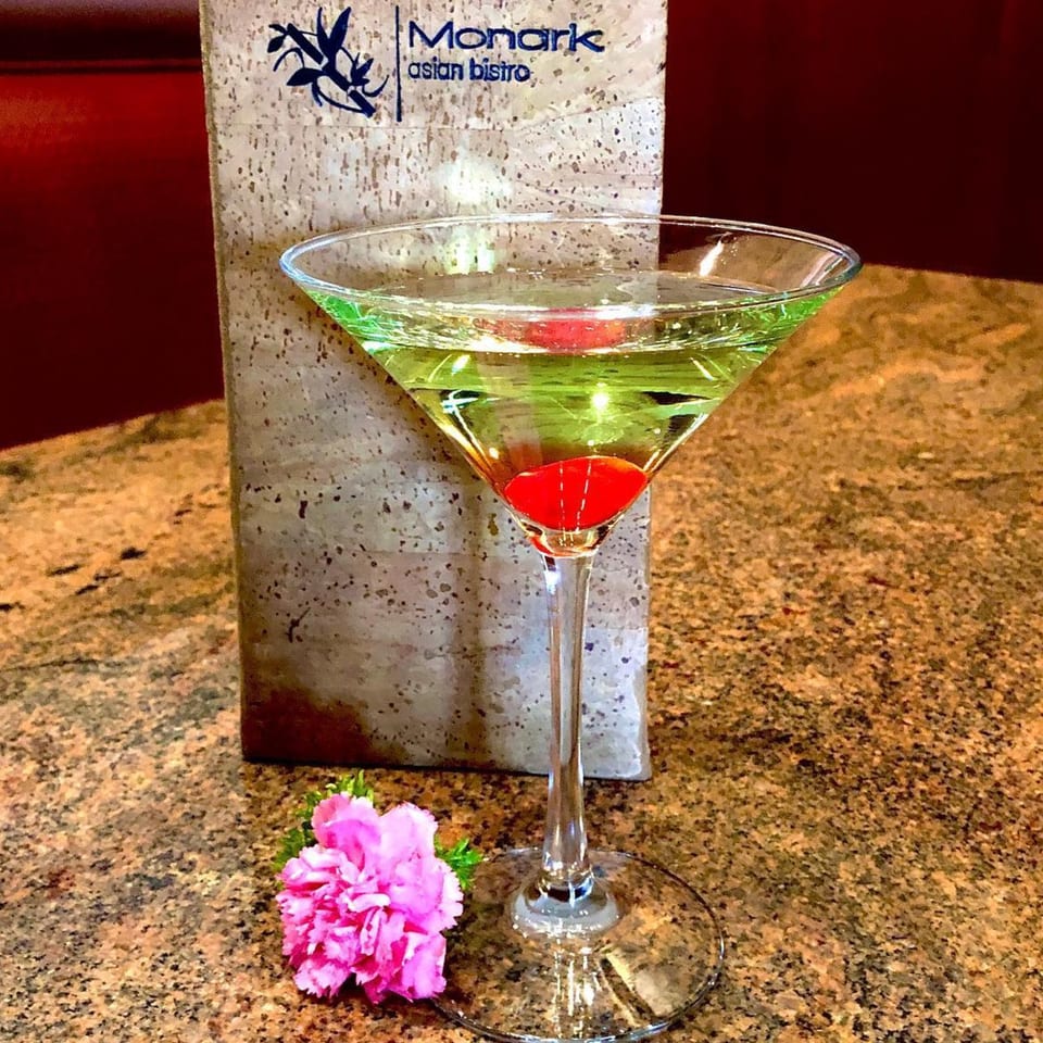 Green martini with a cherry, a pink flower, and a Monark Asian Bistro menu on a granite table.