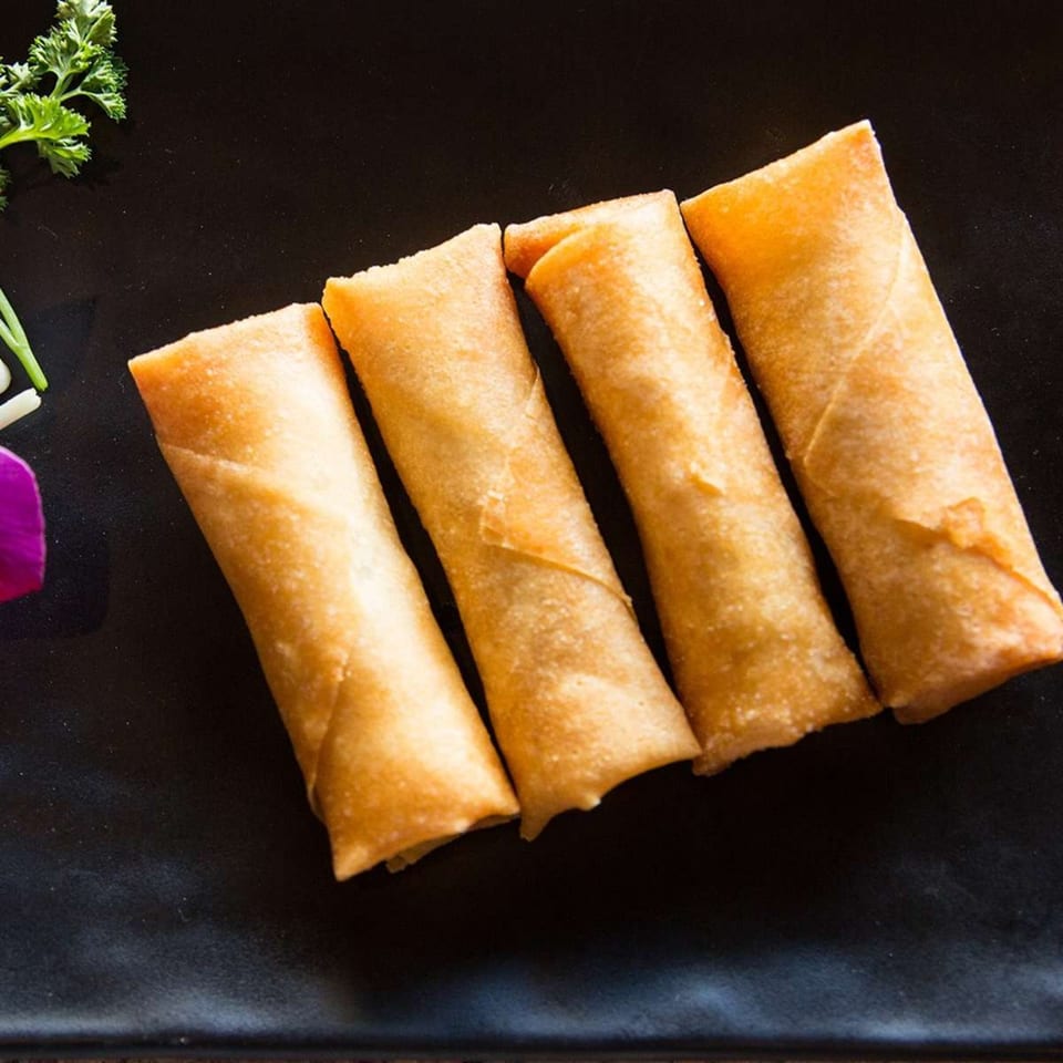 Four golden-brown spring rolls arranged diagonally on a dark plate with green garnish.